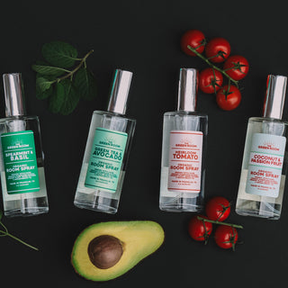 Green Room room and linen sprays in a line on a black background surrounded by ingredients such as plants and botanicals for the Spearmint & Basil, Heirloom Tomato, and Coconut & Passionfruit room & linen sprays