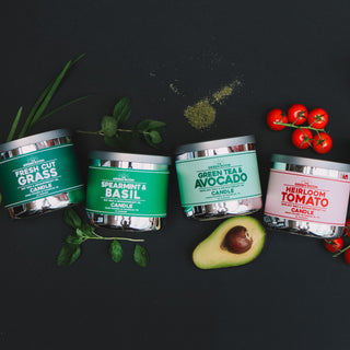 Green Room candles in a line on a black background surrounded by ingredients for the Fresh Cut Grass, Spearmint & Basil, and Heirloom Tomato candles