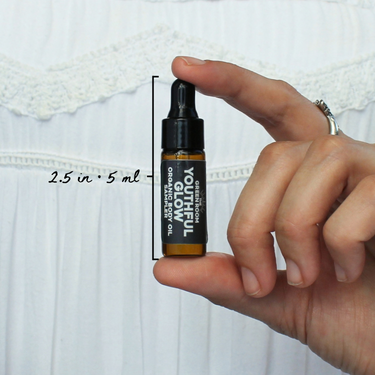 Youthful Glow Organic Body Oil Sampler by The Green Room: a 5 ml amber glass dropper vial held between two fingertips against white lace, marked 2.5 in and 5 ml for scale.