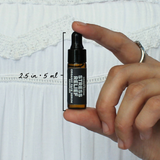 Stress Relief Organic Face Serum Sampler by The Green Room: a 5 ml amber glass dropper vial held between two fingertips against white lace, marked 2.5 in and 5 ml for scale.