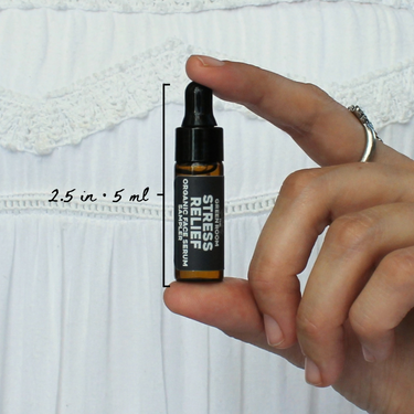 Stress Relief Organic Face Serum Sampler by The Green Room: a 5 ml amber glass dropper vial held between two fingertips against white lace, marked 2.5 in and 5 ml for scale.