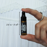 Stress Relief Organic Body Oil Sampler by The Green Room: a 5 ml amber glass dropper vial held between two fingertips against white lace, marked 2.5 in and 5 ml for scale.