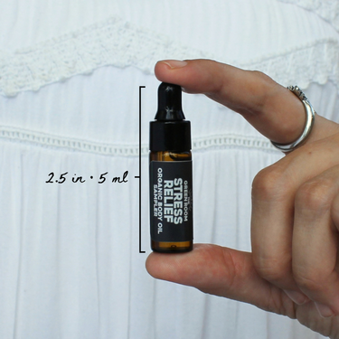 Stress Relief Organic Body Oil Sampler by The Green Room: a 5 ml amber glass dropper vial held between two fingertips against white lace, marked 2.5 in and 5 ml for scale.