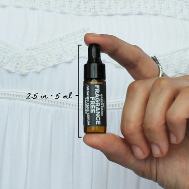 Fragrance Free Organic Face Serum Sampler by The Green Room: a 5 ml amber glass dropper vial held between two fingertips against white lace, marked 2.5 in and 5 ml for scale.