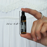 Fragrance Free Organic Body Oil Sampler by The Green Room: a 5 ml amber glass dropper vial held between two fingertips against white lace, marked 2.5 in and 5 ml for scale.