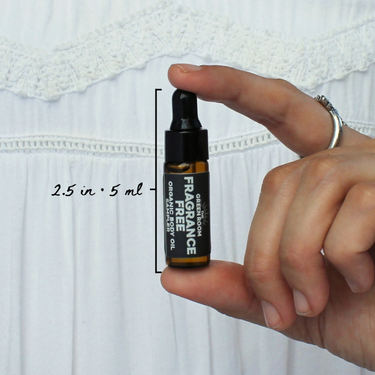 Fragrance Free Organic Body Oil Sampler by The Green Room: a 5 ml amber glass dropper vial held between two fingertips against white lace, marked 2.5 in and 5 ml for scale.