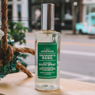 A bottle of Green Room Spearmint & Basil Organic Room Spray stands on a wooden ledge outdoors, with a blurred street scene in the background.