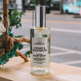 A bottle of Green Room Cedar & Teakwood Organic Room Spray stands on a wooden ledge outdoors with a blurred city street background.