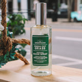 A bottle of Green Room Fresh Cut Grass Organic Room Spray sits on a wooden ledge outdoors with a blurred street scene in the background.