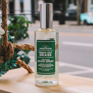 A bottle of Green Room Fresh Cut Grass Organic Room Spray sits on a wooden ledge outdoors with a blurred street scene in the background.
