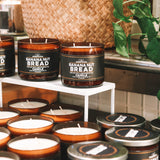 Banana Nut Bread scented candles displayed on white shelves, featuring amber glass jars and metal tins with black labels, set against a background with a woven basket and green plant leaves.