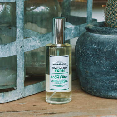 A bottle of Great Green Farm Fern Room Spray stands on a rustic wooden surface, styled with a metal caddy holding glass bottles, a potted cactus, and a dark green background.