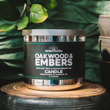 A jar candle labeled 'Green Room Oakwood & Embers' sits on a round wooden pedestal, with dark ceramic bowls and green leaves in the background.