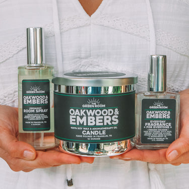 A woman in a white eyelet top holds three Green Mountain Candle products: a South Spray bottle, a Oakwood & Embers Candle tin, and a Preservative bottle.