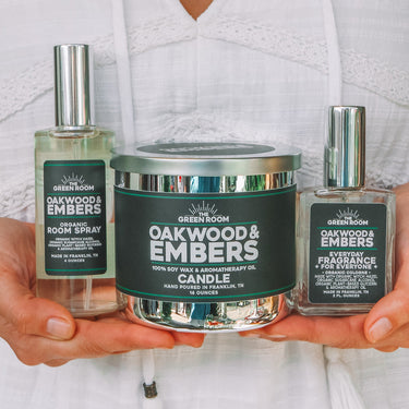 A woman in a white eyelet top holds three Green Mountain Candle products: a South Spray bottle, a Oakwood & Embers Candle tin, and a Preservative bottle.
