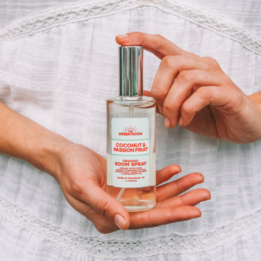 A person in a white lace-trimmed dress holds a clear glass bottle of Green Room Coconut & Passion Fruit room spray. The person's hands and torso are visible against the white fabric background.