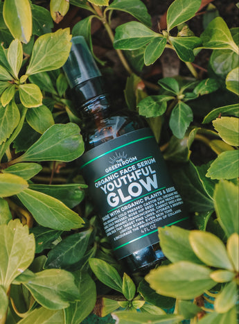 The Green Room Youthful Glow organic face serum in an amber bottle, resting among green leaves.