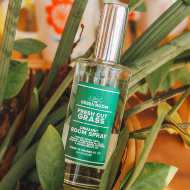 A bottle of Green Room Fresh Cut Grass Organic Room Spray with a silver cap, nestled among green leaves and plant stems.