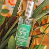 A bottle of Greenroom Spearmint & Basil Organic Room Spray placed among green leaves and soil, with a warm, softly blurred background.