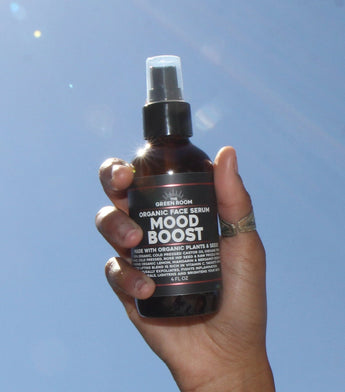 The Green Room Mood Boost organic face serum in an amber glass bottle, held up against a blue sky crested by a light leak.
