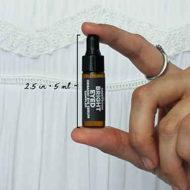 Bright Eyed Organic Eye Serum Sampler by The Green Room: a 5 ml amber glass dropper vial held between two fingertips against white lace, marked 2.5 in and 5 ml for scale.