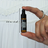 Mood Boost Organic Face Serum Sampler by The Green Room: a 5 ml amber glass dropper vial held between two fingertips against white lace, marked 2.5 in and 5 ml for scale.
