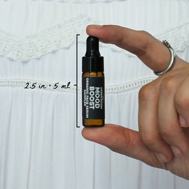 Mood Boost Organic Face Serum Sampler by The Green Room: a 5 ml amber glass dropper vial held between two fingertips against white lace, marked 2.5 in and 5 ml for scale.
