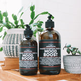 Two bottles of Mood Boost Organic Everyday Body Oil with labels indicating the product is organic, cold pressed, with organic plants, and made in Franklin TN. One bottle is larger and the other is smaller, both with black caps and placed on a wooden surface with a green potted plant in the background.