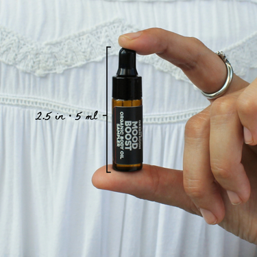 Mood Boost Organic Body Oil Sampler by The Green Room: a 5 ml amber glass dropper vial held between two fingertips against white lace, marked 2.5 in and 5 ml for scale.