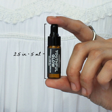Youthful Glow Organic Face Serum Sampler by The Green Room: a 5 ml amber glass dropper vial held between two fingertips against white lace, marked 2.5 in and 5 ml for scale.