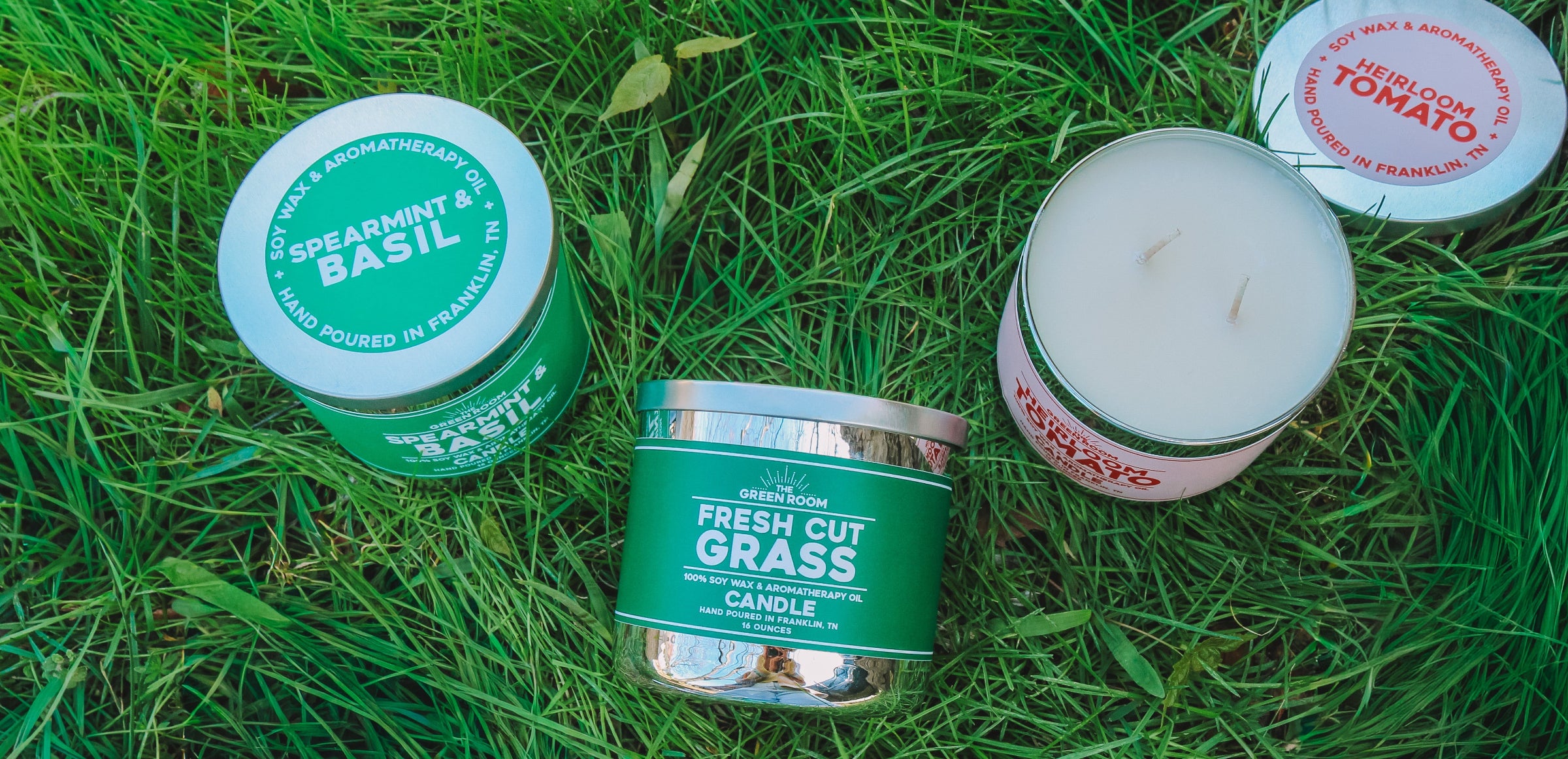 Three 100% soy wax candles lying in fresh grass: Spearmint & Basil, Fresh Cut Grass, and Heirloom Tomato (opened to show  double cotton wicks)