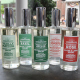 Five Green Room scented spray bottles lined up on a dark tabletop, with a blurred street and storefront background and a floral mug at the right edge.