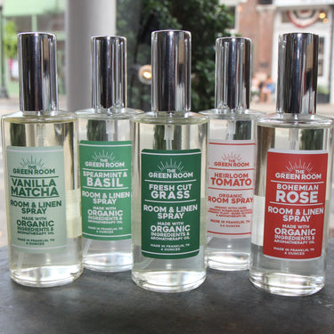 Five Green Room scented spray bottles lined up on a dark tabletop, with a blurred street and storefront background and a floral mug at the right edge.