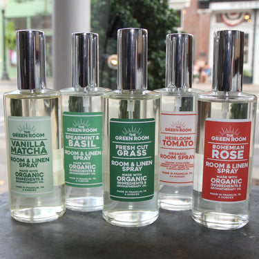 Five Green Room scented spray bottles lined up on a dark tabletop, with a blurred street and storefront background and a floral mug at the right edge.