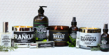 The Green Room product lineup on white hexagon tile: Cedar & Teakwood cologne, Franklin 100% soy wax candle, Youthful Glow organic body oil, Oakwood & Embers roll-on cologne, a lit Banana Nut Bread candle, Fragrance Free face serum, Palo Santo & Saffron candle, Bright Eyed organic eye serum, and 5 ml fragrance samplers.