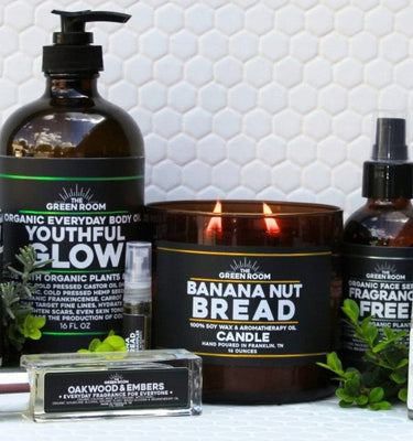 A collection of products by The Green Room on white hexagon tile: Youthful Glow organic body oil in an amber 16 fl oz pump bottle, an Oakwood & Embers roll-on cologne, a lit Banana Nut Bread 16 oz 100% soy wax candle, a 5 ml fragrance sampler, and an organic Fragrance Free face serum.